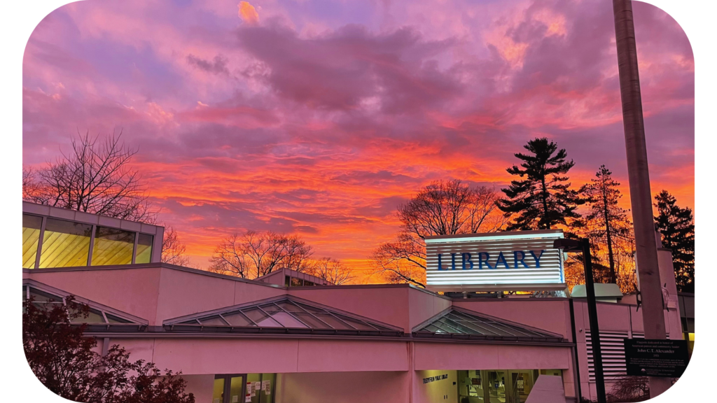 Tredyffrin Public Library shown at sunset. The sunset has deep yellows, oranges, pinks, and light purples. The LIBRARY sign atop the building is lit.